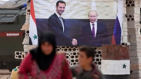 A Syrian woman walks with a boy past a banner showing Russian President Vladimir Putin shaking hands with Syrian President Bashar al-Assad, June 1, 2018 