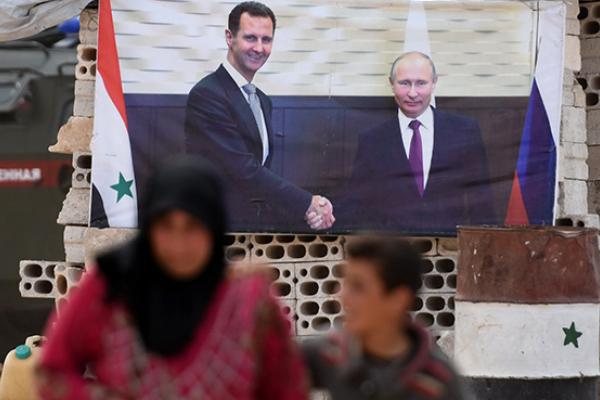A Syrian woman walks with a boy past a banner showing Russian President Vladimir Putin shaking hands with Syrian President Bashar al-Assad, June 1, 2018 